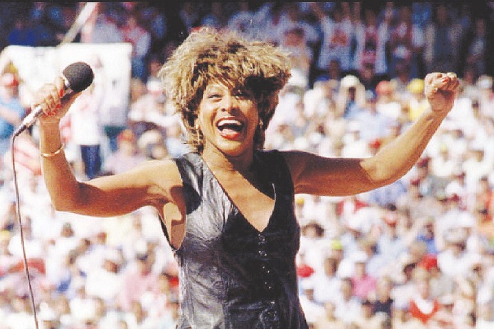Performer singing energetically on stage with a microphone, wearing a leather outfit and smiling.