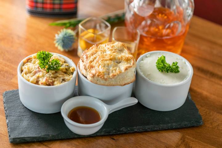 Three dishes on slate: scrambled mixture, pie, mashed potatoes with garnish; small sauce cup and drinks behind.