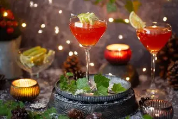 Two red cocktails with garnish, surrounded by candles and greenery in a festive setting.